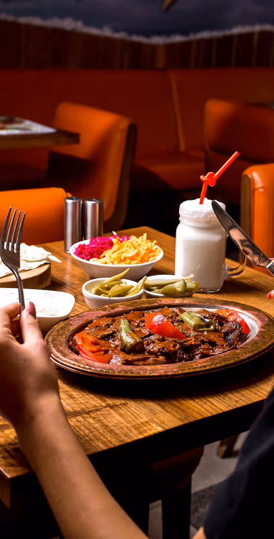 Person holding fork and knife ready to eat a plated meat dish garnished with vegetables, with pickles, salad bowls, and a milkshake on a wooden table in a restaurant.